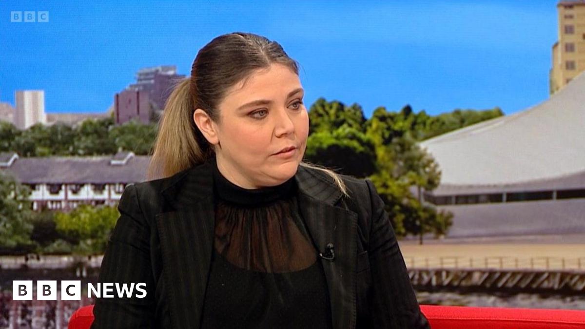 A woman with long brown hair and wearing a black top and black suit jacket is sitting on the red BBC Breakfast sofa. Images of Media City in Salford can be seen behind her.