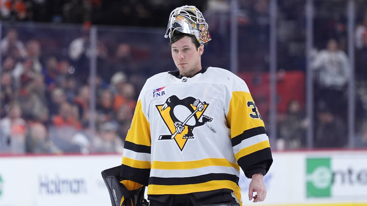 A white man is wearing white-gold-and-black hockey uniform. He is a goalie, wearing black-and-yellow leg pads. His mask sits on top of his head, revealing his face.