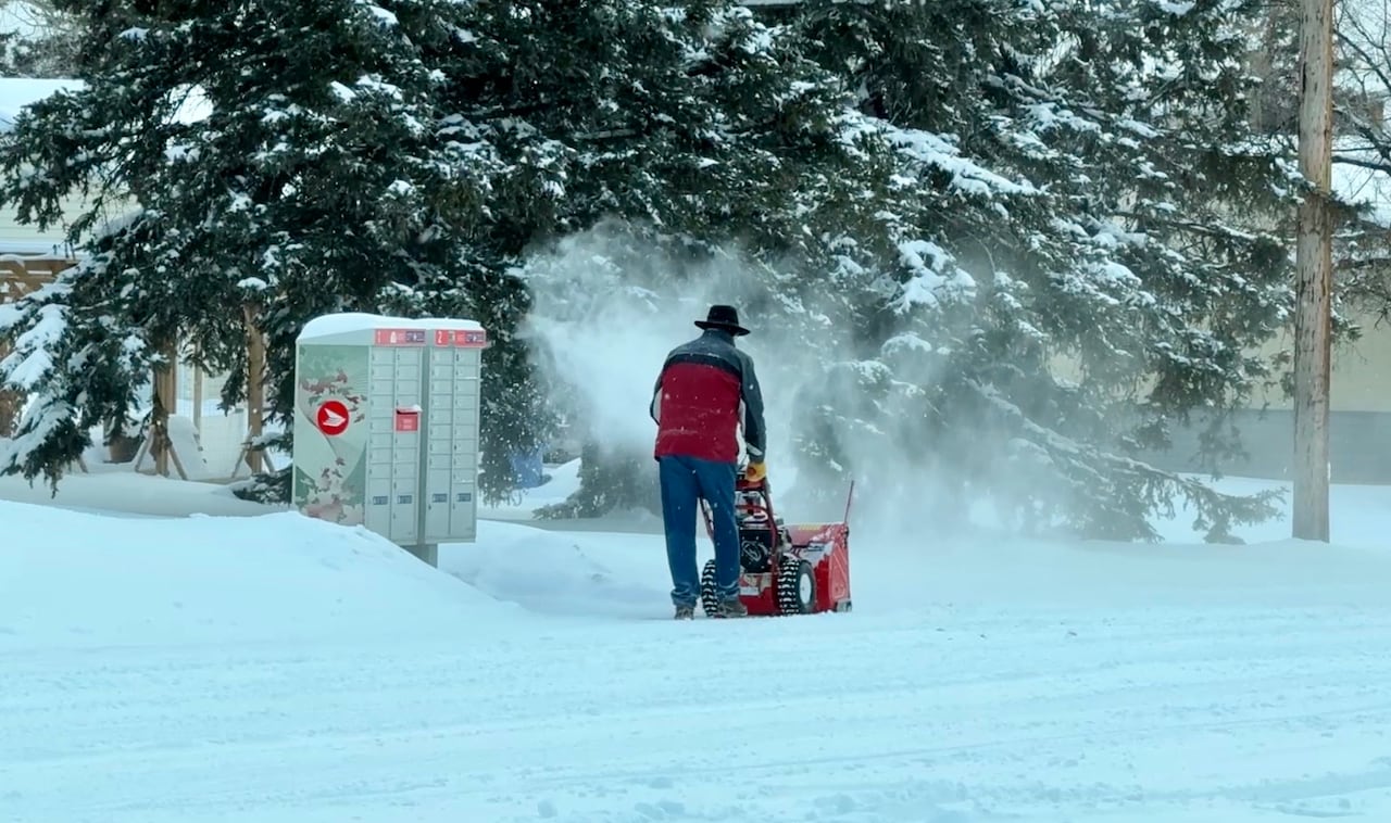 a man in a red and grey coat and cowboy hat pushes a snowblower