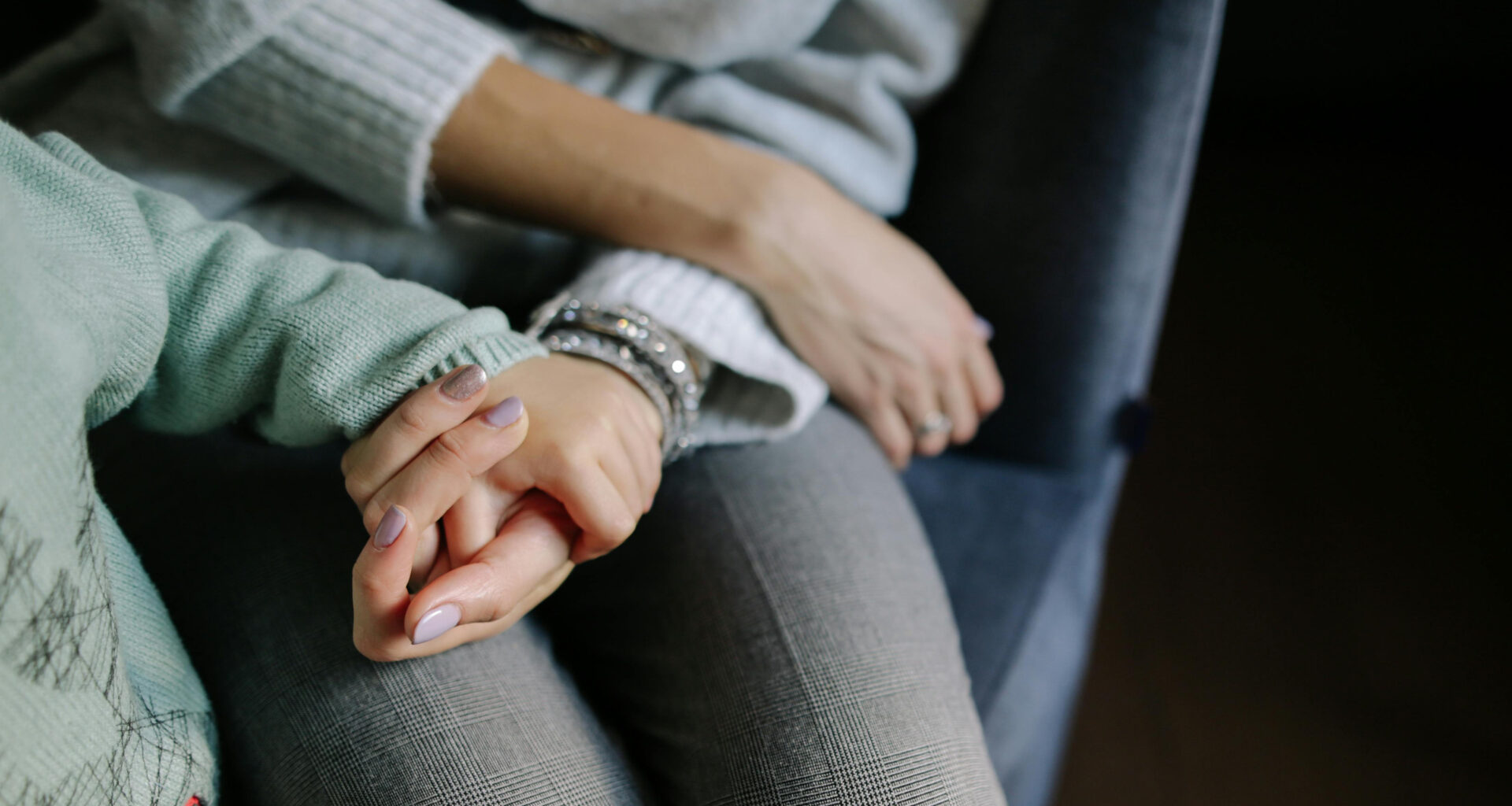 A woman’s hand cupped upwards holding a toddler’s hand with both rested on the woman’s leg.