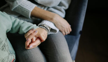 A woman’s hand cupped upwards holding a toddler’s hand with both rested on the woman’s leg.