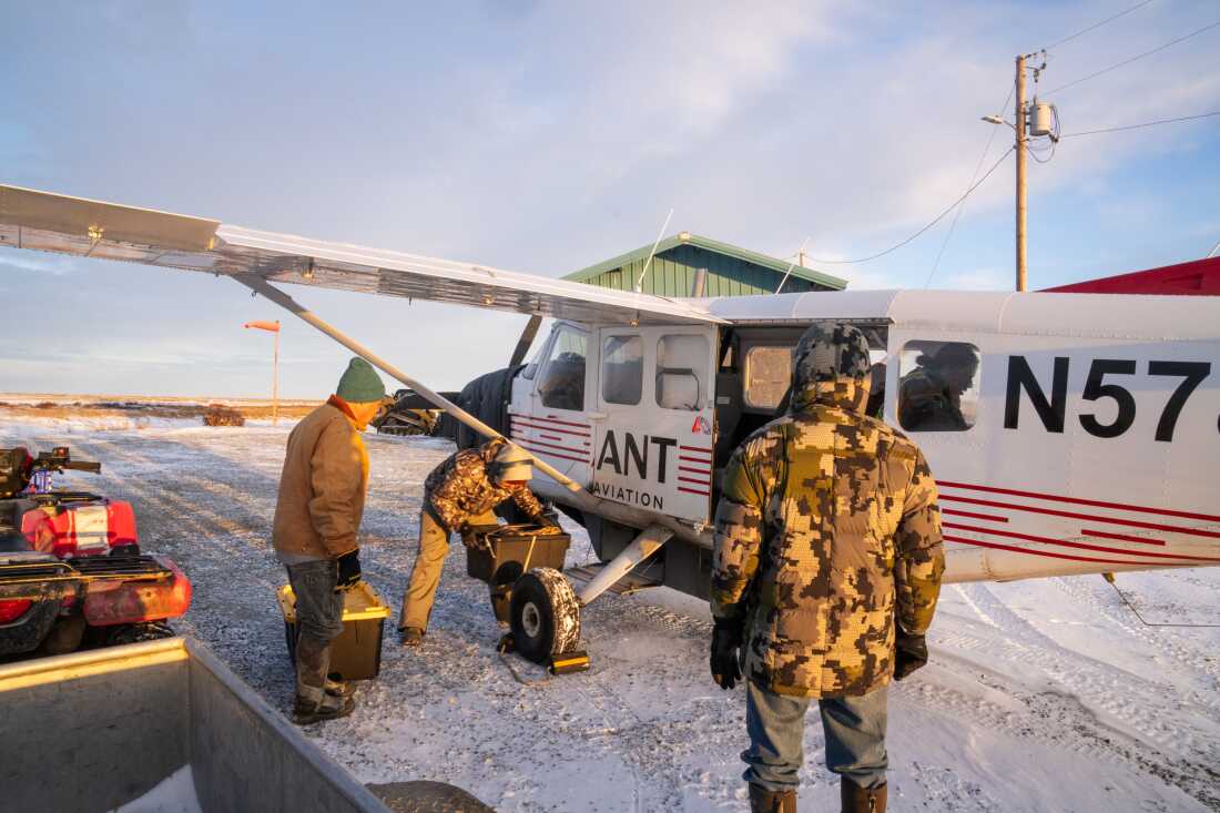 Three Kwigillingok men wearing winter coasts load plastic bins of belongings into a small plane that's parked on snowy ground.
