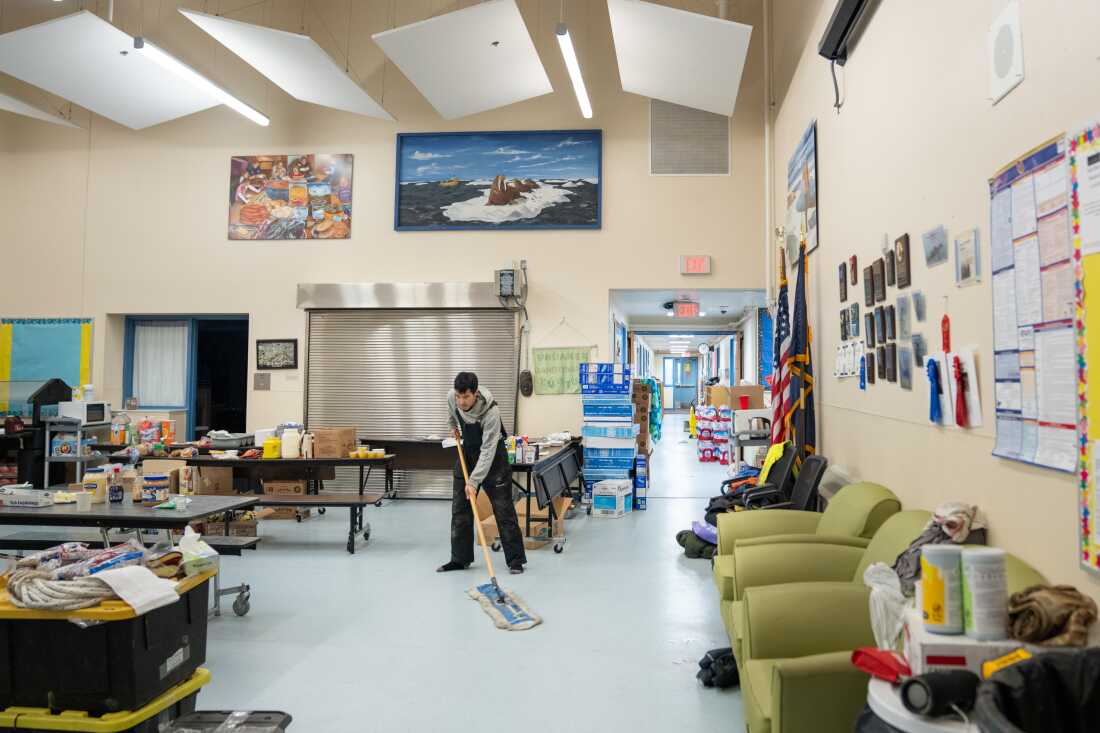 A man mops a floor in a large gathering room at the Kwigillingok School. Groceries are on some long tables, and bottled water is stacked in a hallway that leads out from the back of the room.