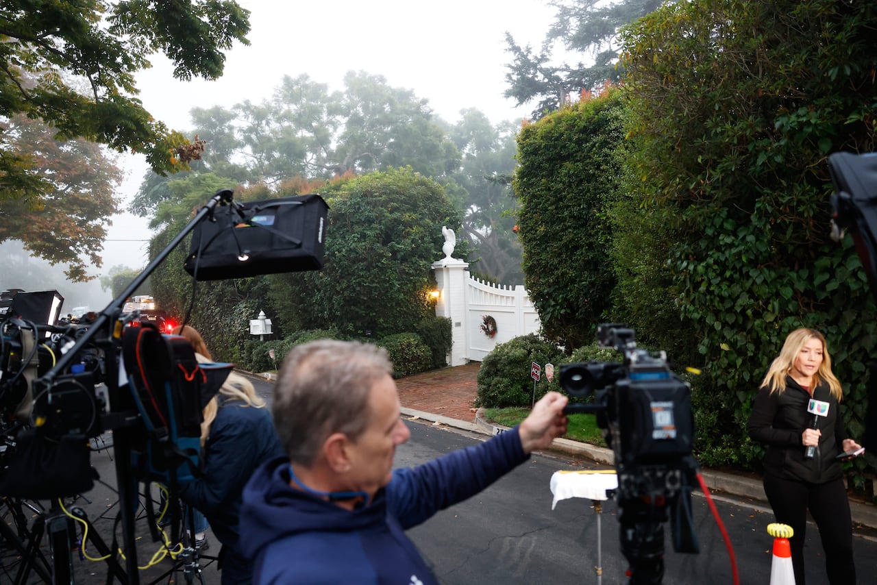 In the foreground, a man adjusts a standup camera in front of a gate between trees and shrubs.