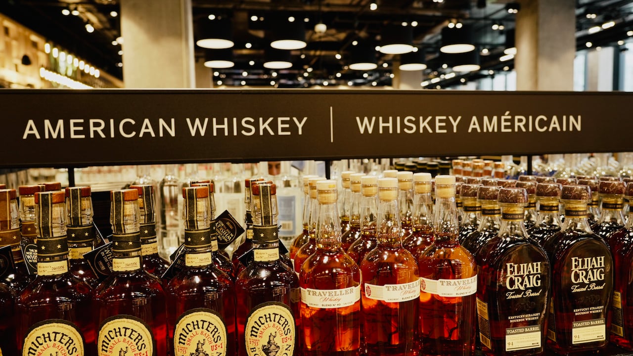 Rows of bottles sit on a shelf in front of a sign reading 'American Whiskey.' 