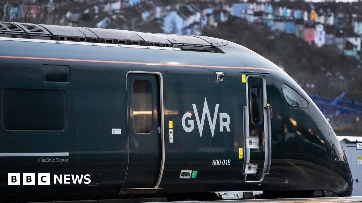 Image of a GWR train pulling into Bristol station. The dark green train takes up most of the image, with the Totterdown houses seen blurred in the background.
