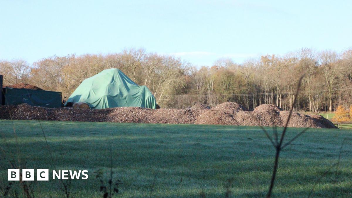A large green tarpaulin covers a mound of materials on a field, with several piles of brown waste nearby. Trees line the background.
