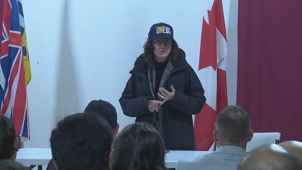 A woman in a 'OneBC' hat speaks to an audience in front of the B.C. and Canadian flags.