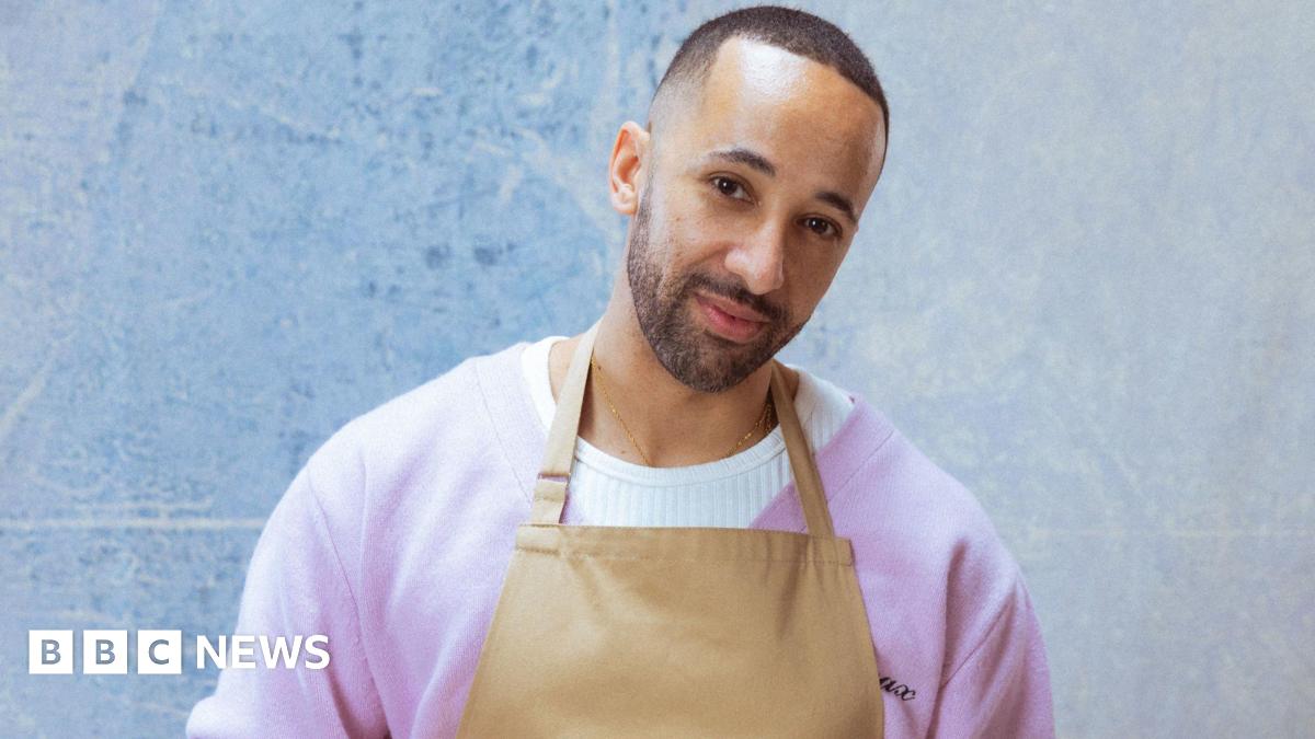 A man with short black hair and beard stands with his head cocked to one side. He is wearing a purple cardigan over a white jumper, with a brown apron over the top.