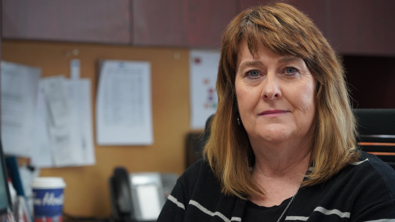 A woman with brown hair, wearing a black sweater, sits in an office.