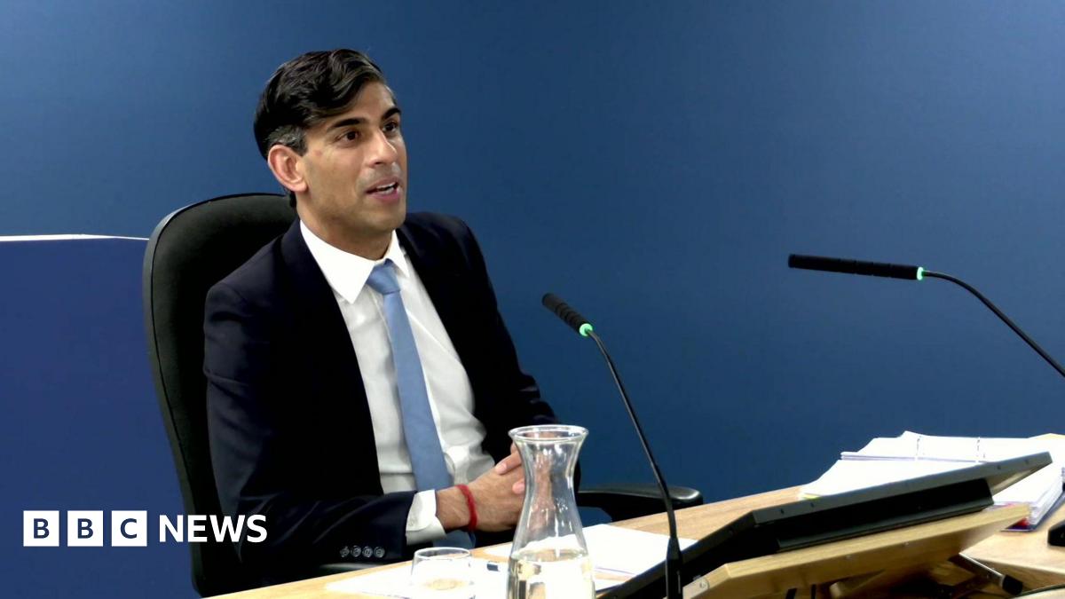 Former Prime Minister Rishi Sunak, wearing a blue suit, white shirt and light blue tie, sits at a desk with microphones and papers in front of him