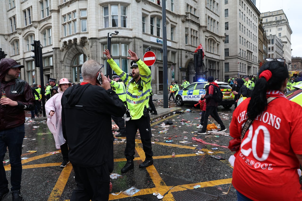A bearded police officer in reflective vest stands in the middle of an intersection and appears to yellow toward several people in the area.