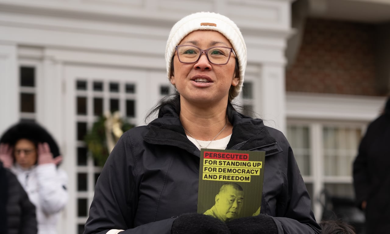 woman in jacket speaking at a rally and holding a pamphlet written Persecuted for standing up for democracy and freedom