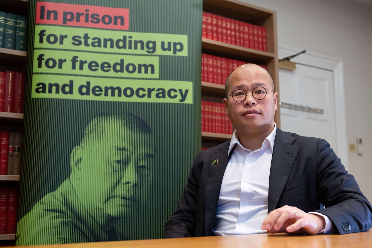 man with glasses sitting next to poster written in prison for standing up for freedom and democracy
