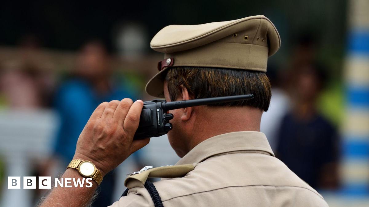 Indian police in brown uniform and cap with back to camera holding walkie talkie