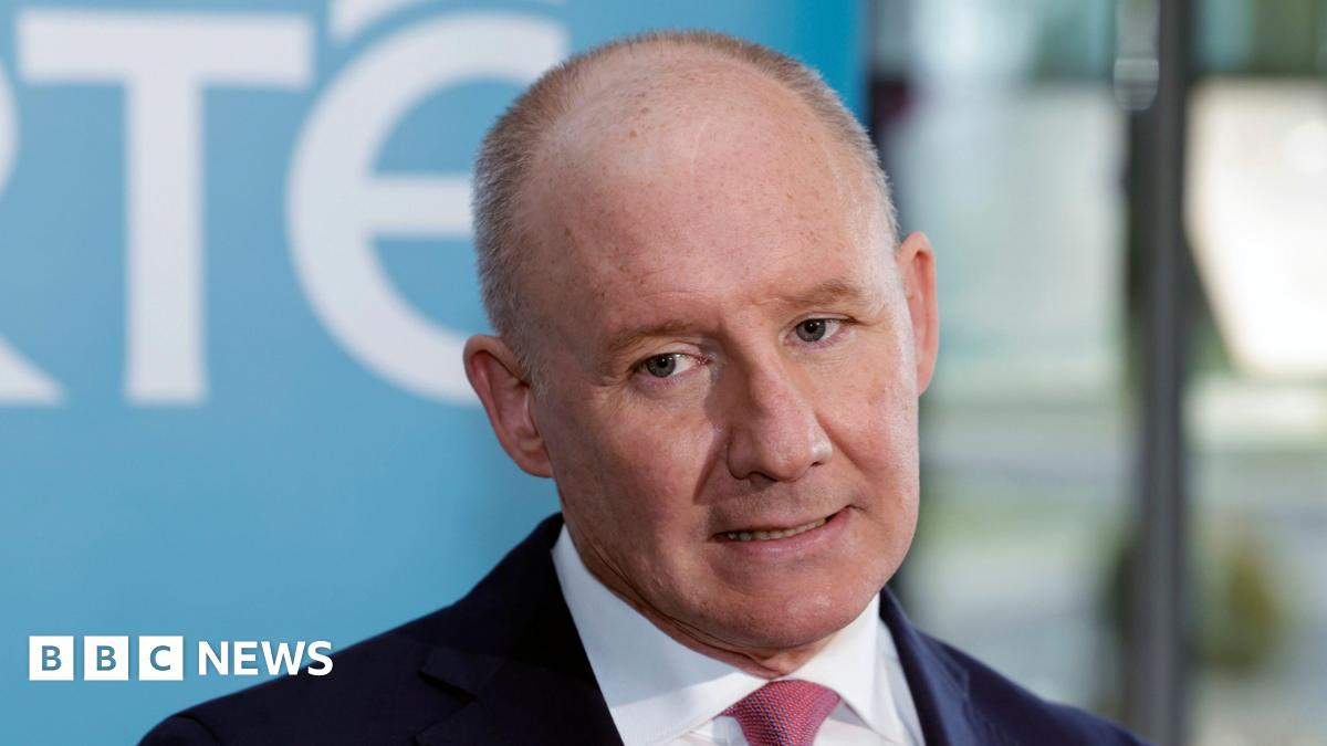 Jim Gavin, a man with thinning grey hair, is wearing a navy suit and red pattern tie. The background is out of focus but the blue and white of an RTÉ banner can be seen.