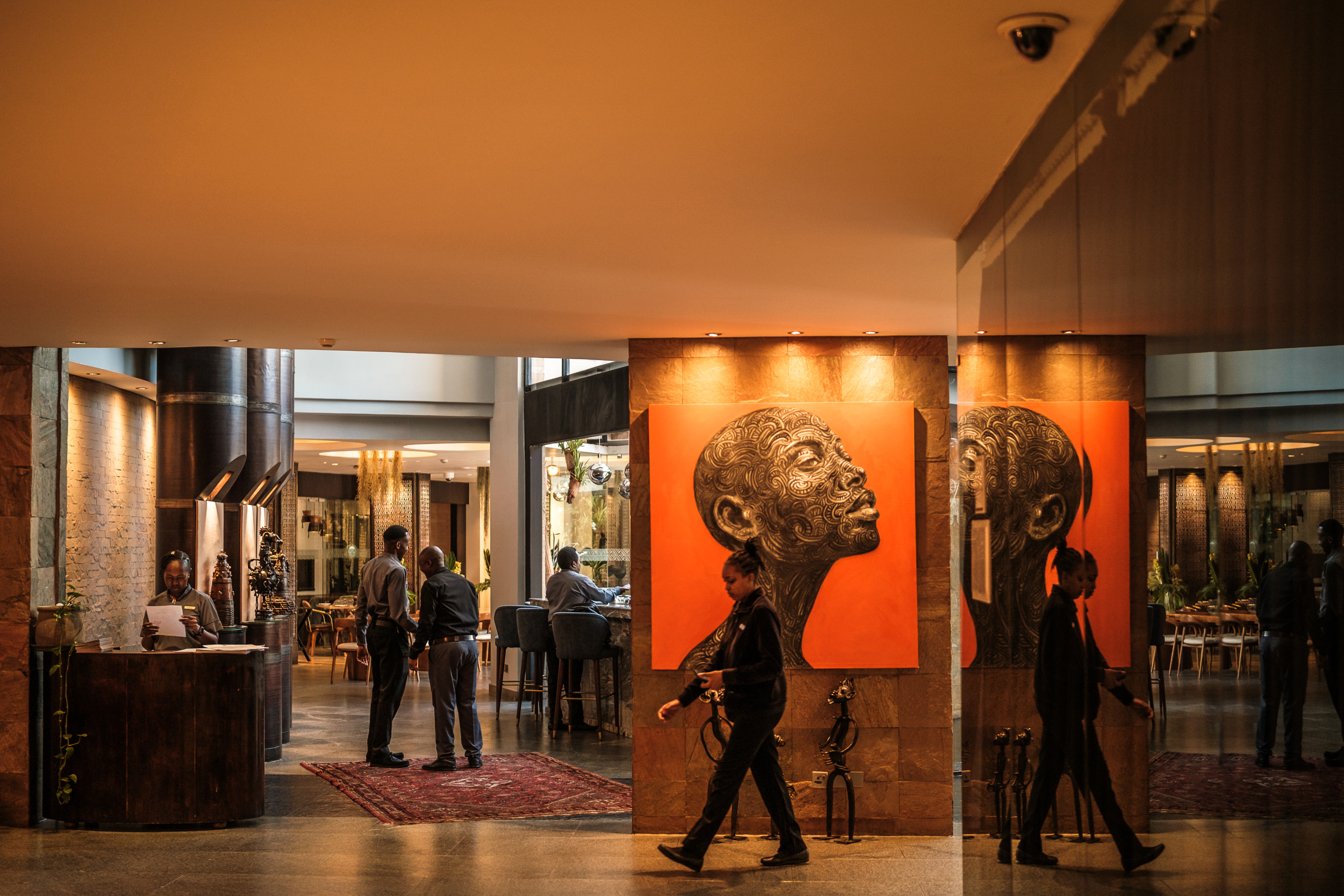 A fancy hotel lobby and front desk. Several employees go about their day. The lobby is decorated with African artwork, including a large painting of a person’s face.