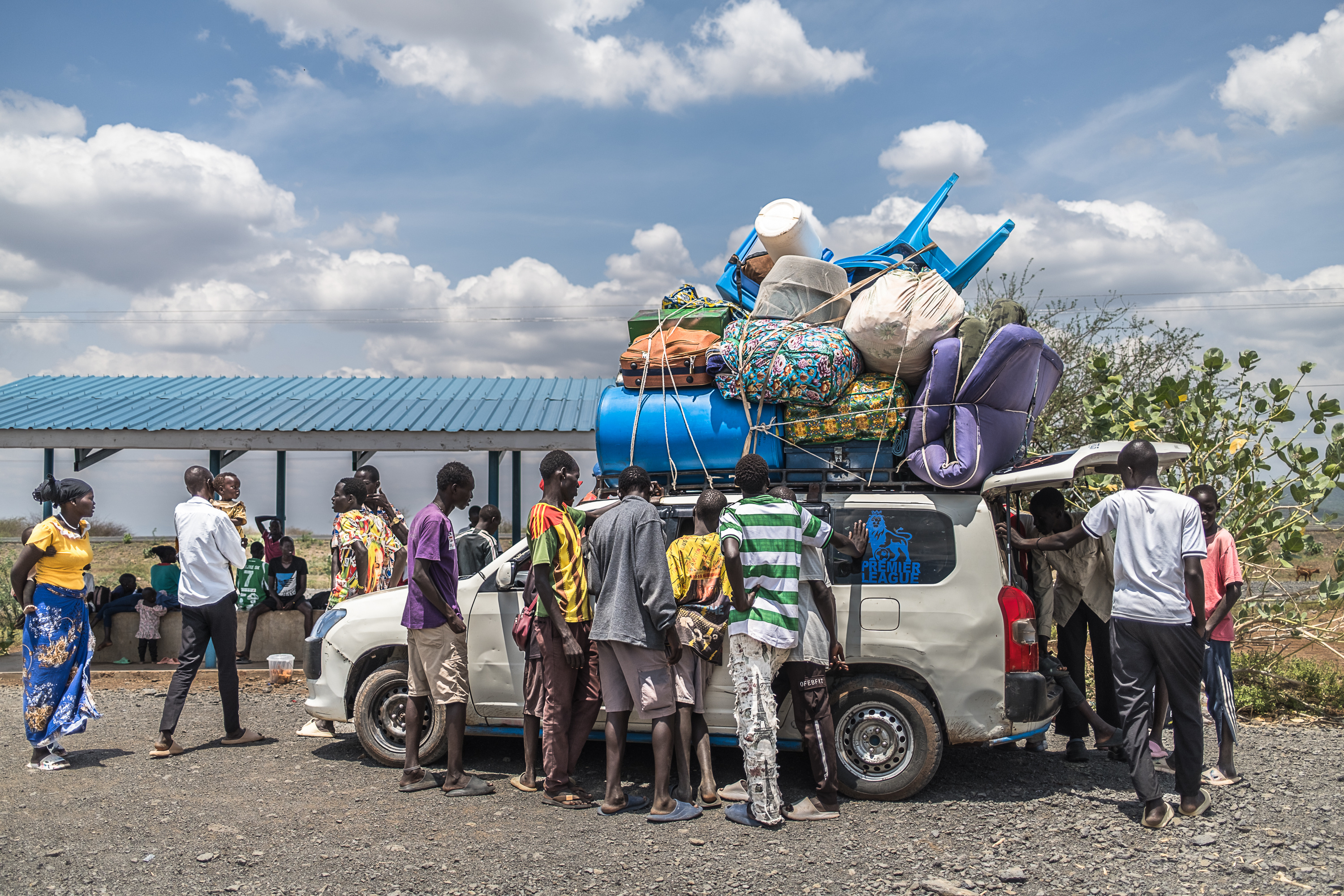 People gather around a hatchback car. Several of them are climbing in the back even though the car is already full. Luggage and furniture are piled high on top of the car and tied in place.