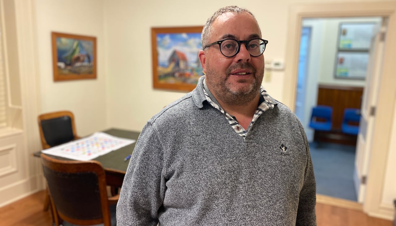 A white man with glasses wearing a grey sweater stands in an office with paintings on the wall behind him, and a door open to the hallway