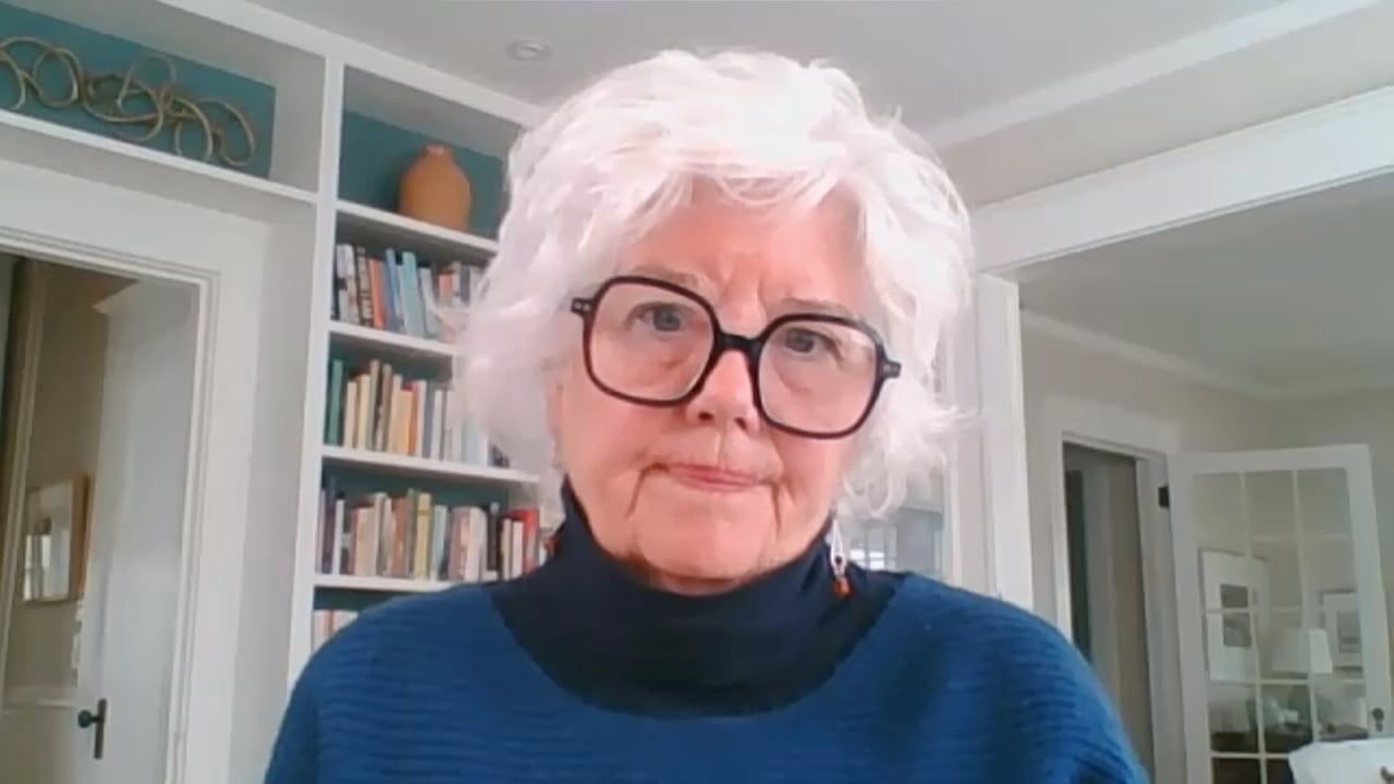 A white woman with short white hair and glasses wears a blue sweater. Doorways and a bookshelf are behind her.