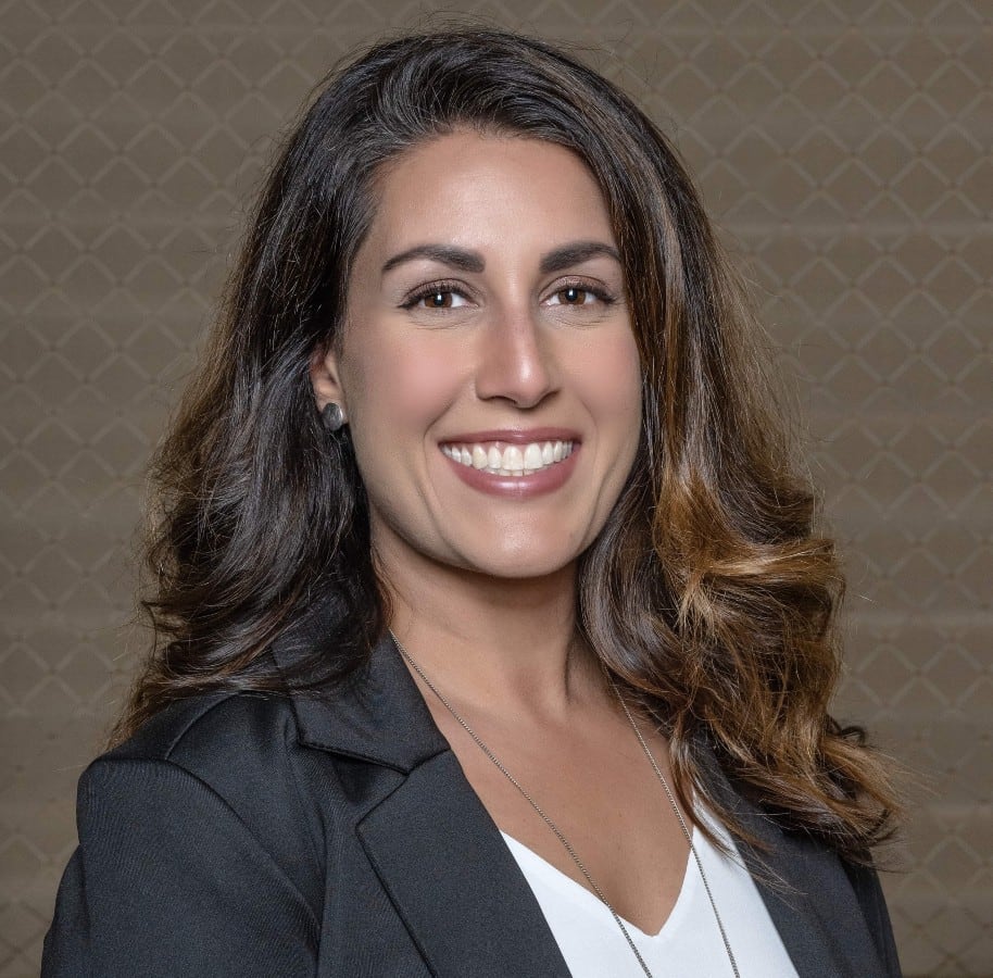 A professional headshot of a smiling white woman with long brown hair in a grey blazer