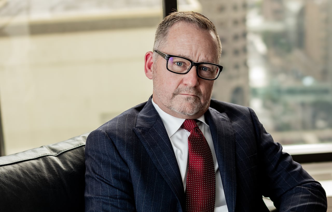 A man in a suit sits in a chair and looks at the camera.