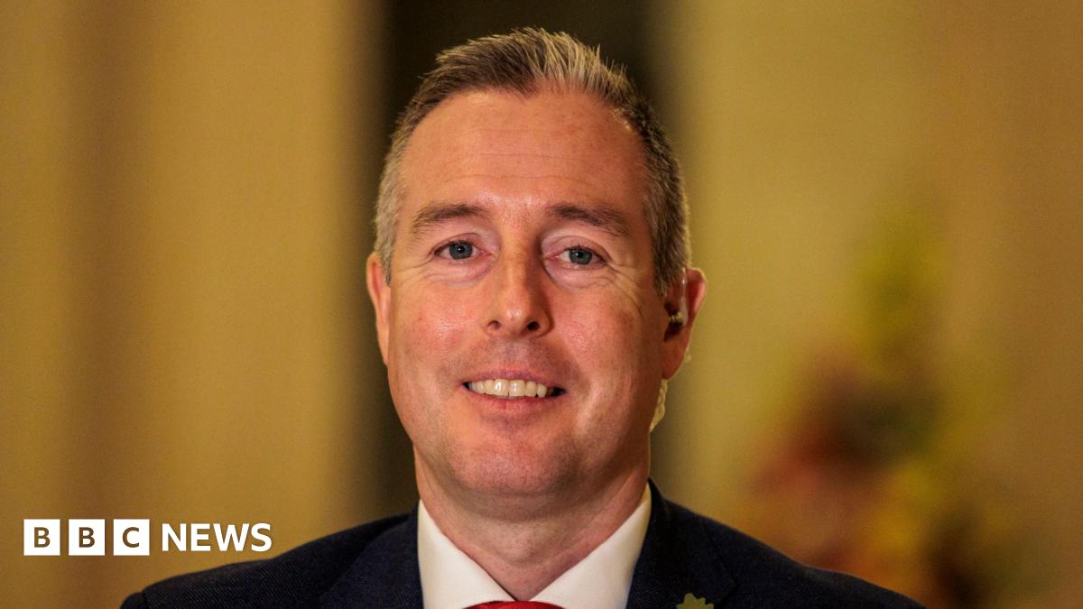 Paul Givan inside Stormont, he is wearing a navy suit and a white shirt with a red tie. He has short white hair.