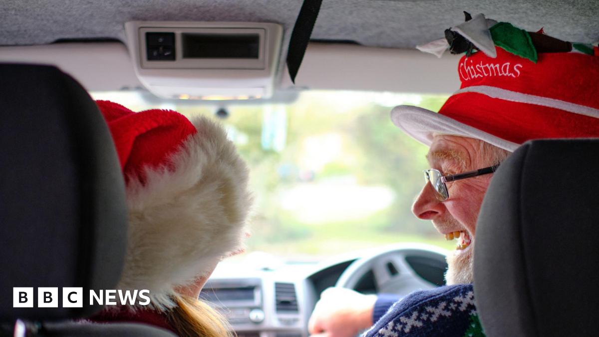 A man and a woman sitting in a car. Both are wearing Christmas hats. The man is driving and smiling at the woman