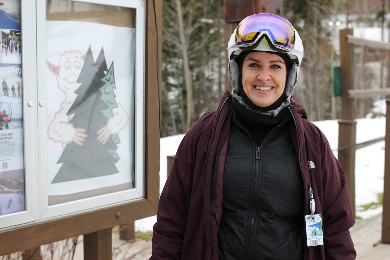 A woman in a black vest and purple ski jacket poses for a photo at a ski hill.