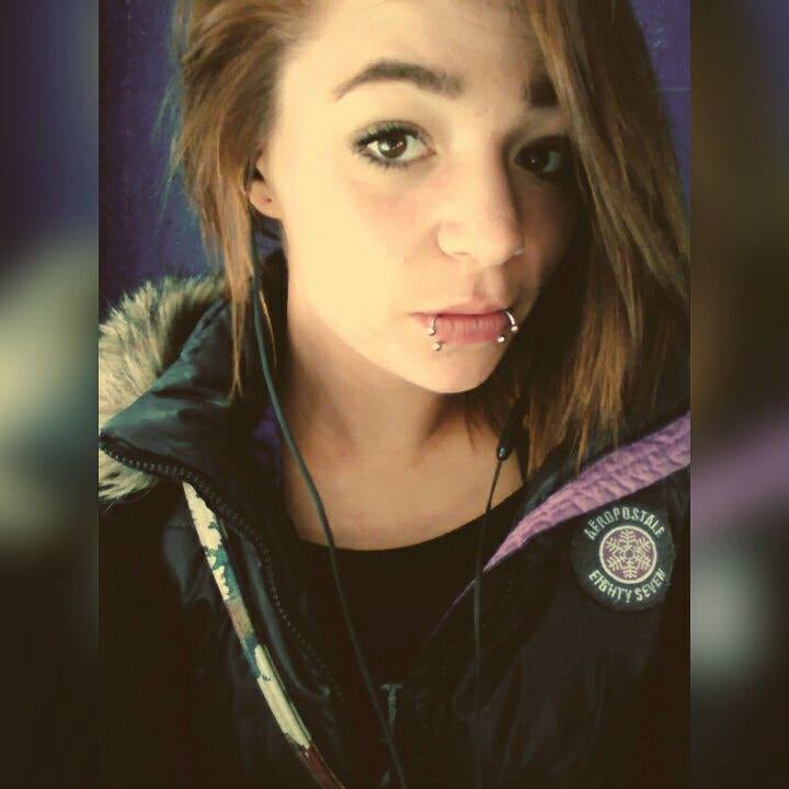 A portrait image of a young Caucasian woman with shoulder-length brown hair, brown eyes. She has two lip piercings, hoops with baubles. 