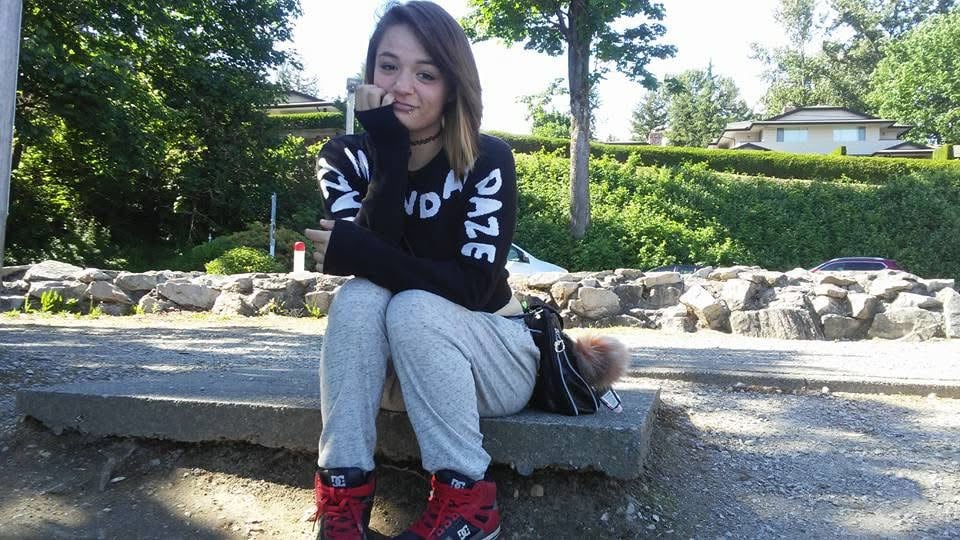A girl with medium length hair sits on a curb smiling with her head on her hand.