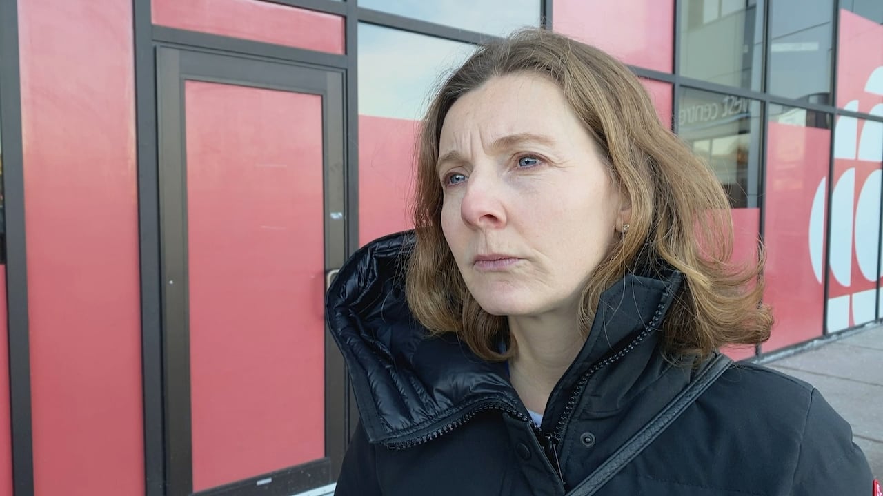 A white woman with light brown hair stands outside in a black winter jacket, with red windows of a building behind her