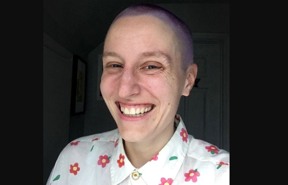 A white woman with a shaved head smiles into the camera. She wears a white floral shirt.