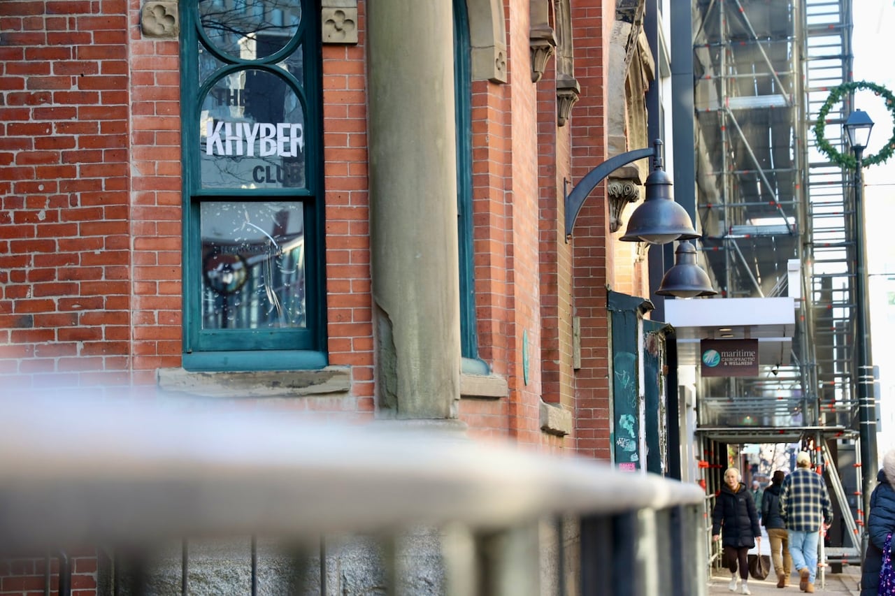 A close-up view of the side of a red brick building, with The Khyber Club seen in a window as people walk by