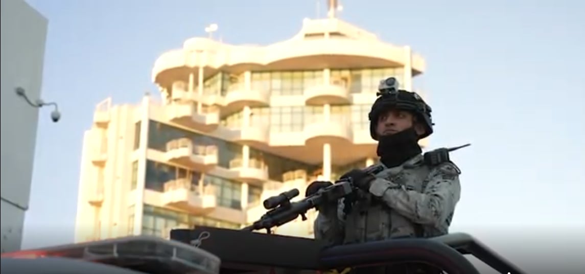 A man with a gun and a helmet with an apartment building in the background.