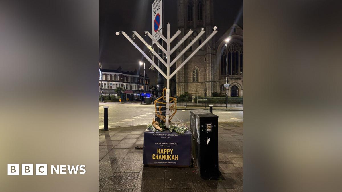 A large decorative silver Hanukkah menorah on a street, on a stand which reads 'Happy Chanukah'. The lightbulbs representing candles on the top of the menorah have been damaged.