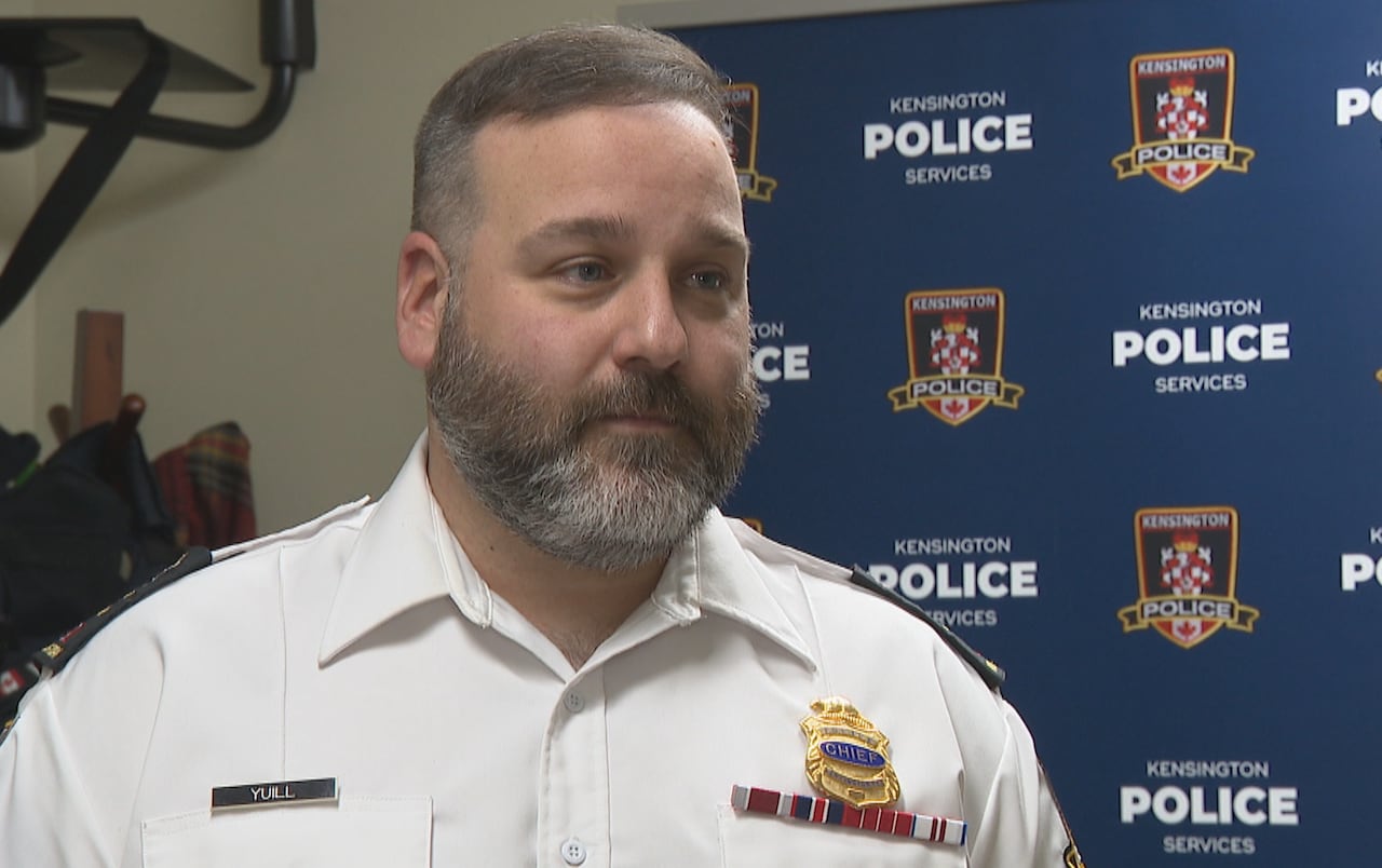 A medium close-up of Kensington Police Chief wearing a white uniform with a name tag and badge, speaking in front of a blue Kensington Police Services backdrop.