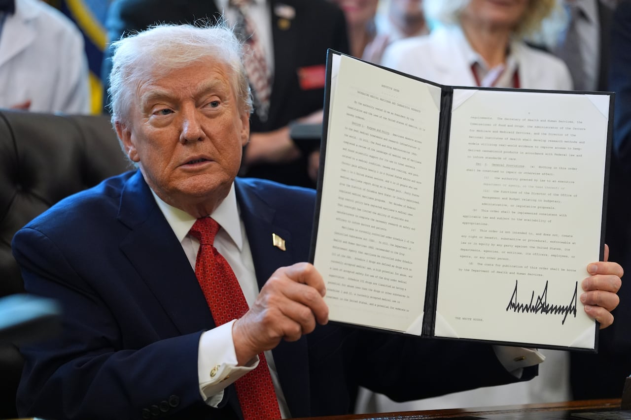 Donald Trump, wearing a navy blue suit, white shirt and red tie, sits behind a desk and holding up a folder with his signature on a document.