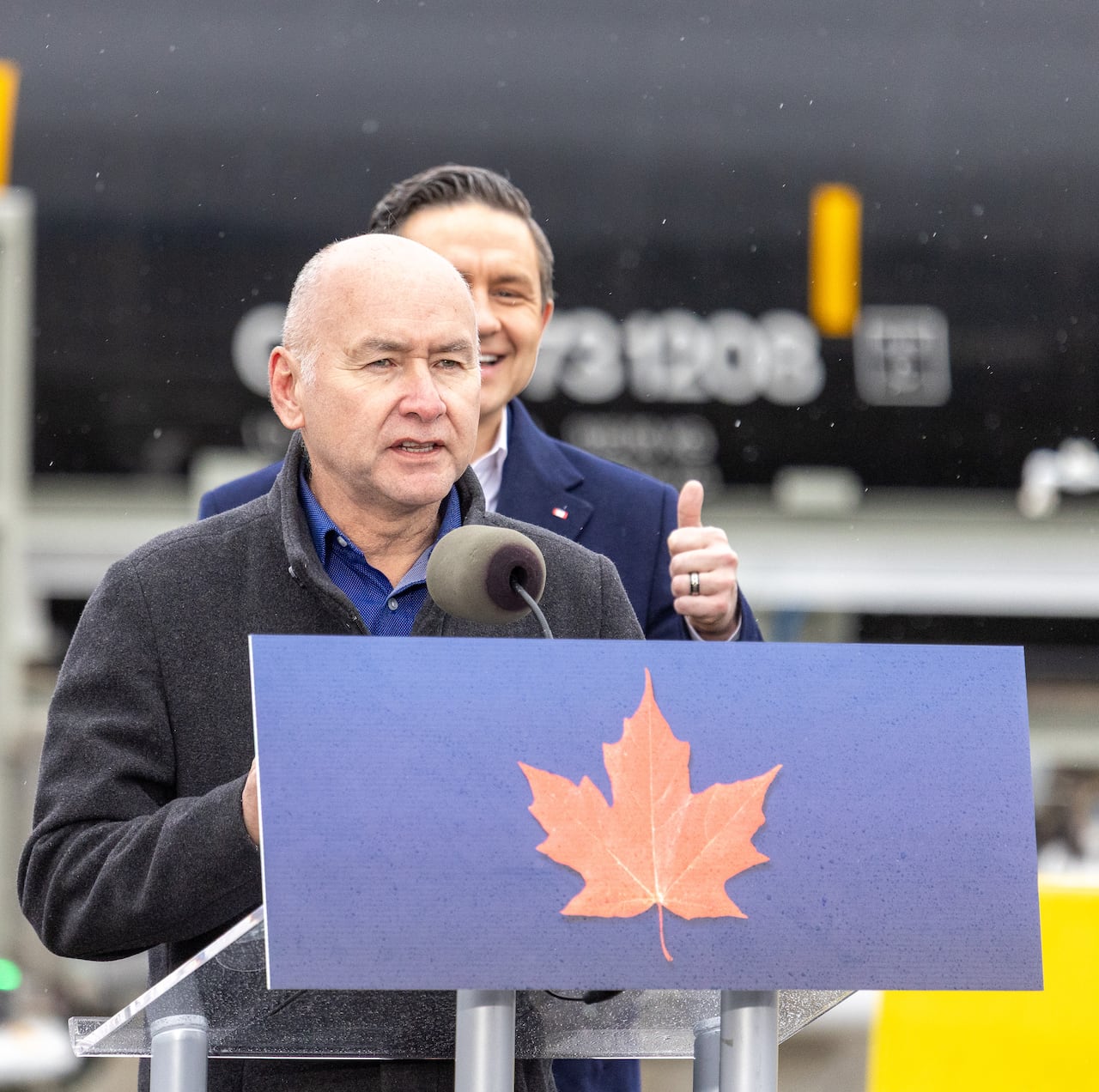 A bald man speaks at a podium while another man behind him gives a thumbs up.