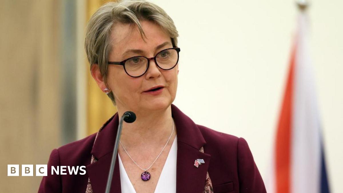 Yvette Cooper, who has cropped blonde hair and wears dark-framed glasses, speaks at a lectern in front of a British flag