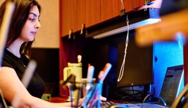 A professional with long hair focuses on a computer screen, seated at a cluttered wooden desk with a laptop and various office supplies in the foreground.