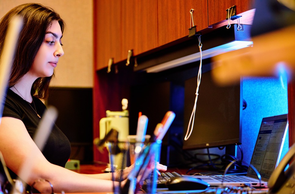 A professional with long hair focuses on a computer screen, seated at a cluttered wooden desk with a laptop and various office supplies in the foreground.
