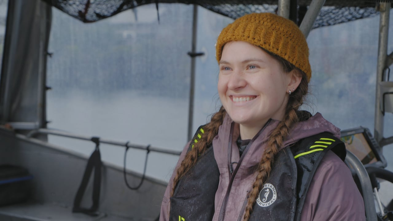 A woman in rain gear and a life jacket smiles in front of a boat