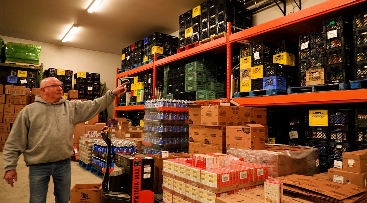 Man in grey sweater points to shelves of crates and boxes.