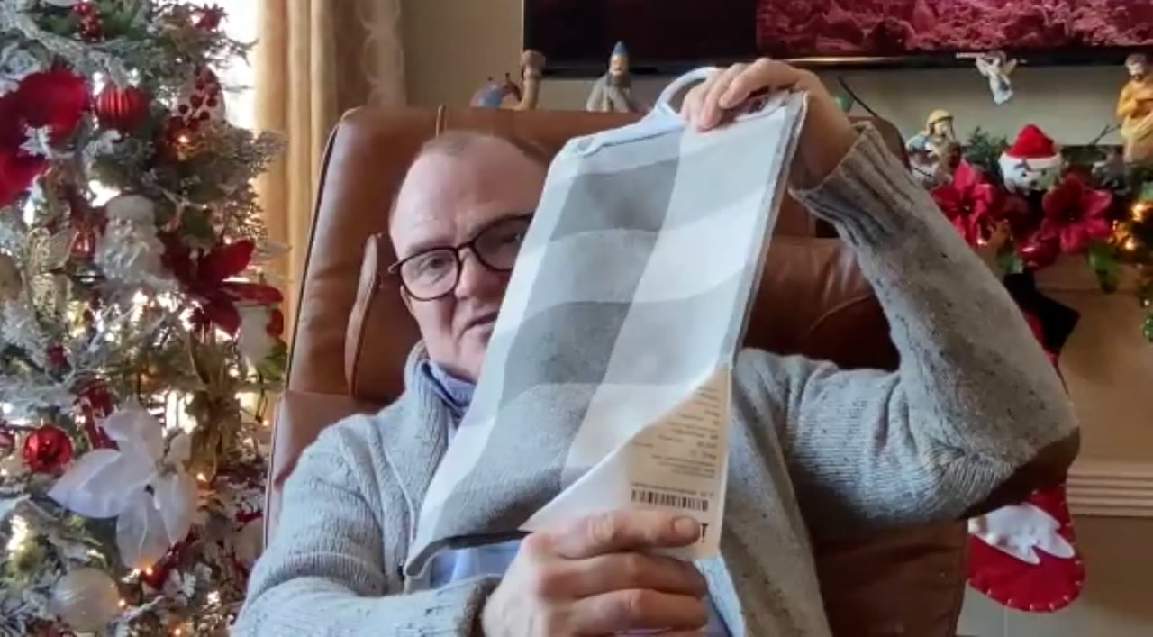 Man in grey sweater holds up cloth surrounded by Christmas decorations.