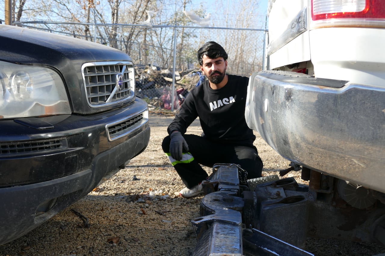 A man is crouching down between the bumpers of two vehicles. He is outside.