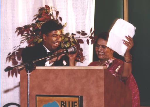 Two people stand at a podium. One is holding up a piece of paper in their left hand.