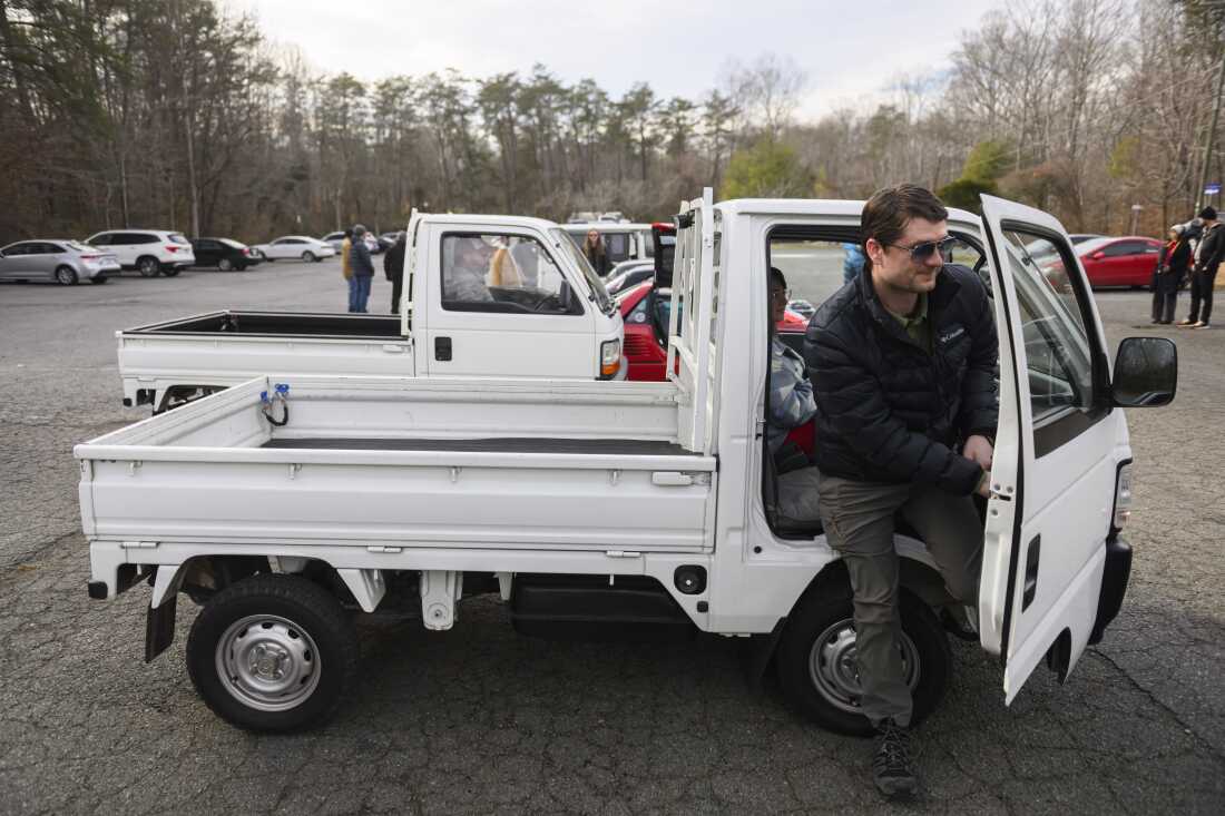 Ryan Douglass steps out of the right door of his white Honda Acty kei pickup, which is parked in a parking lot.