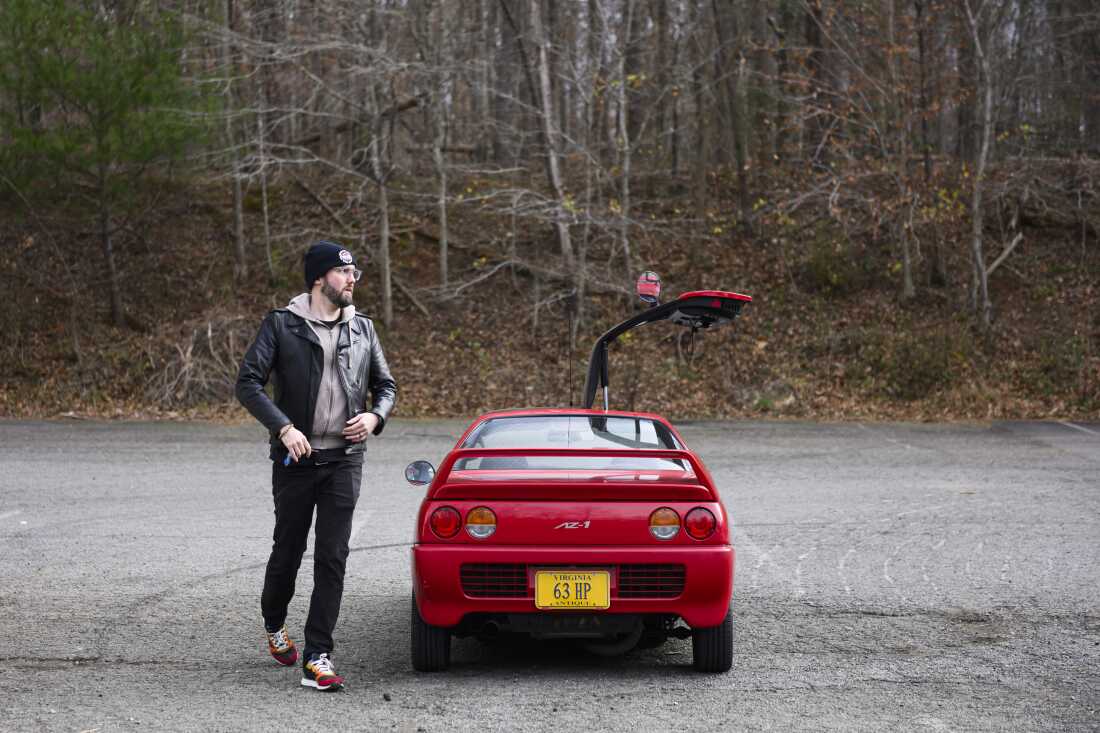 On the left side of the frame, Andrew Maxon walks by his red Autozam AZ-1, whose right gull-wing door is raised. The back of the two-door kei car faces the camera.