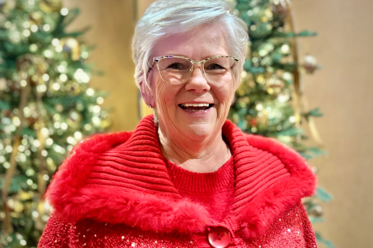 A woman with grey hair and glasses smiles, wearing a red sweater.